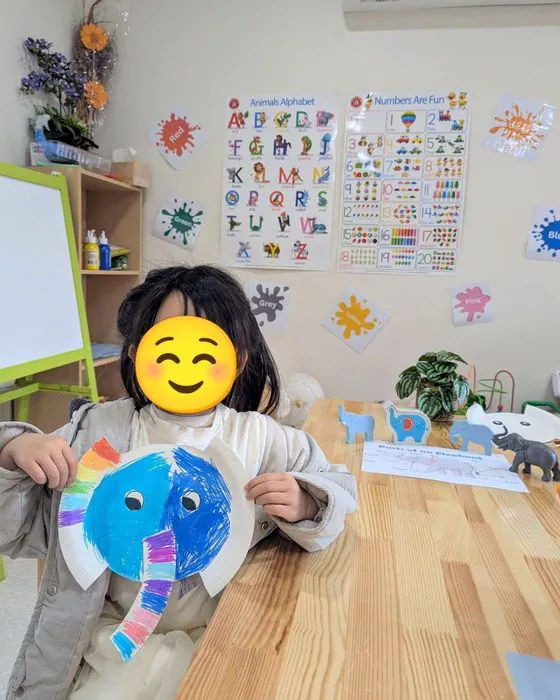 A child proudly holding up a painted paper plate elephant